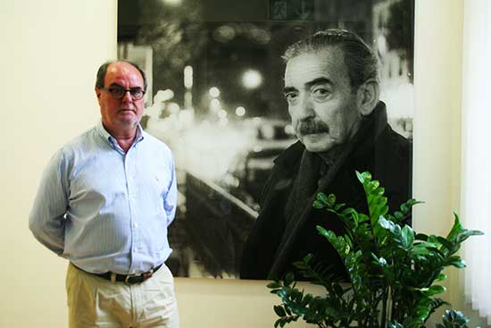 "Rafael de la Dueña, bibliotecario de la Biblioteca Juan Gelman, junto a la imagen del autor que da nombre a la misma y recibe al usuario de la Biblioteca en el Instituto Cervantes de Viena"