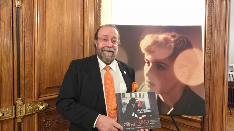 Jussi Brofeldt con la publicación AHO &amp; SOLDAN – Helsinki 1950-luvun varjossa, frente a una imagen de Claire Aho, en la recepción celebrada en Viena. Foto: CulturaLatina.